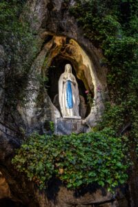 Opplyst Jomfru Maria-statue i en steingrotte, omgitt av grøntområder.