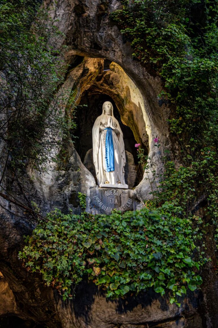 Opplyst Jomfru Maria-statue i en steingrotte, omgitt av grøntområder.