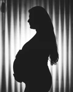 Monokrom silhuett av en gravid kvinne som står i profil mot gardinbakgrunn.