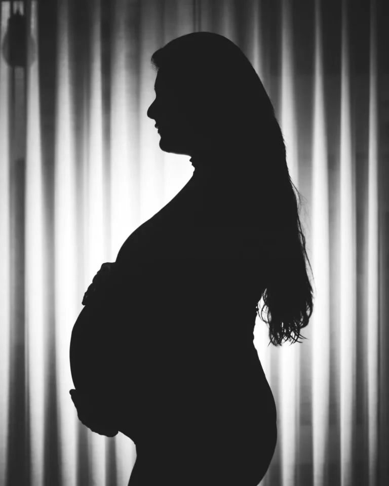 Monokrom silhuett av en gravid kvinne som står i profil mot gardinbakgrunn.